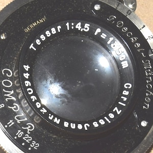 May include: A black Carl Zeiss Jena Tessar 1:4,5 f=13.5cm camera lens with a Compur shutter mechanism. The lens features white engraved text including 'GERMANY', 'Tessar 1:4,5 f=13.5cm', 'Carl Zeiss Jena Nr. 2690944', and 'F. Deckel-München'.