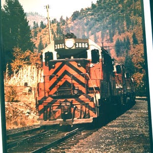 May include: Vintage orange and black train engine with chevron stripes on the front. The train is on a track with a backdrop of trees and a mountain. The train has the number 3006 on the front.
