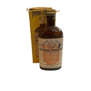 May include: A vintage brown glass bottle with a white label that reads "Phenol Sodique" and a yellow box with the same text. The bottle is a medical product and the label includes information about the product's use and manufacturer.