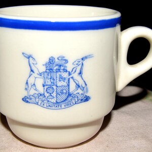 May include: Cream-colored ceramic mug with a blue rim and handle. The mug features a blue emblem with two deer and a crest, and the text "EX UNITATE VIRES". The mug has a stacked base.
