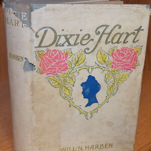 May include: A vintage book titled "Dixie Hart" by Will N. Harben. The cover features a stylised illustration of two pink roses with green leaves and a blue silhouette of a woman's head.