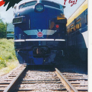 Pode incluir: Uma locomotiva azul e branca com as palavras "Potomac Eagle Train" e "Where Eagles Fly!" no lado. O trem está em uma via com uma área verde gramada no fundo. O texto "West Virginia Wild and Wonderful" está em um adesivo oval verde abaixo do trem.