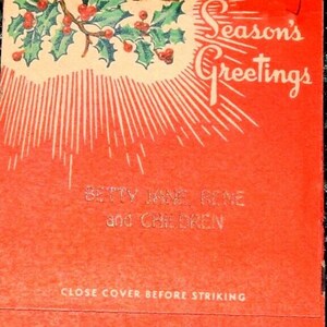 Op de afbeelding: Rode luciferdoosdeksel met een hulsttak en bessen, een zonnestralenontwerp en de tekst "Season's Greetings" in wit. De tekst "Betty Jane Rine and Children" is gedrukt onder het zonnestralenontwerp. De tekst "Close cover before striking" is gedrukt aan de onderkant van de luciferdoosdeksel.