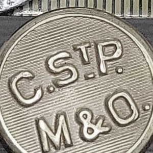 Pode incluir: Um token de metal prateado com o texto "C.S.T.P. M&O." em relevo.