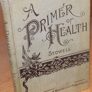 Puede incluir: Un libro antiguo titulado "A Primer of Health" de Stowell, publicado por Silver Burdett & Co. La portada presenta un diseño floral decorativo en marrón y blanco.