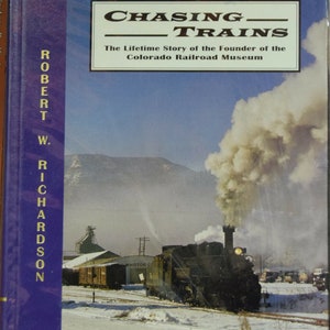 May include: A book cover with a blue background and the title "Chasing Trains: The Lifetime Story of the Founder of the Colorado Railroad Museum" in black text. The cover features a black steam locomotive with white smoke billowing from the chimney, set against a snowy mountain landscape.