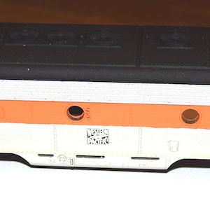 May include: A white and orange model train car with a black roof. The car has a rectangular shape and is made of plastic. The car has a series of small windows on the side and a small door on the side. The car has a black roof with a series of small details on the roof.