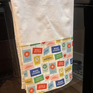 May include: White kitchen towel with a colourful border featuring inspirational phrases like "Make it Better" and "Make Change" in various fonts and colours. The towel is hanging on a black oven.
