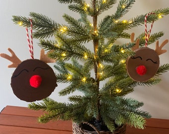 May include: A small artificial Christmas tree decorated with two felt reindeer ornaments. The tree is in a brown wicker basket and has white fairy lights.