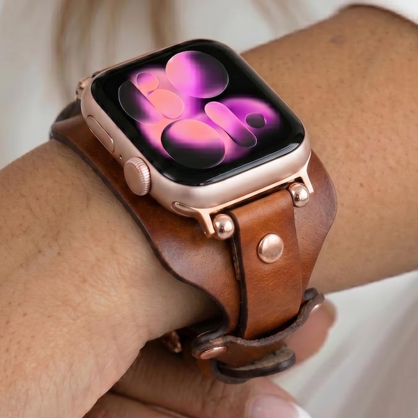 Puede incluir: Un reloj inteligente en oro rosa con una pantalla digital que muestra formas circulares abstractas en rosa y morado. El reloj está sujeto a una correa de cuero marrón ancha tipo brazalete, asegurada con tachuelas metálicas y una hebilla.