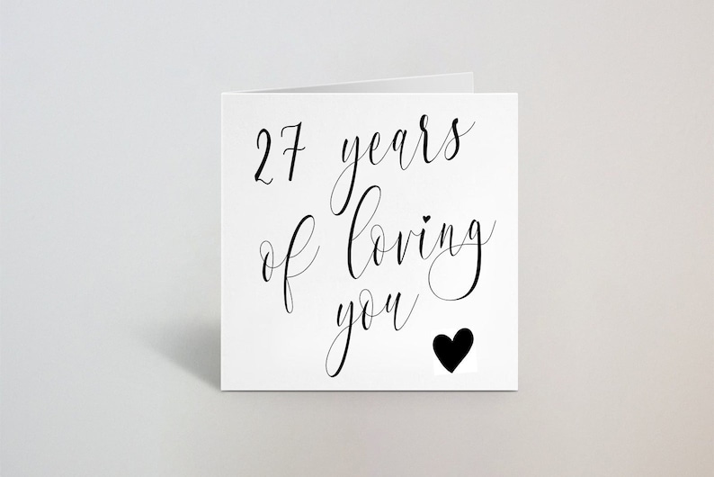 May include: White greeting card with the text "27 years of Loving you" in black script. A black heart is at the bottom right. The card is folded and set against a light gray background.
