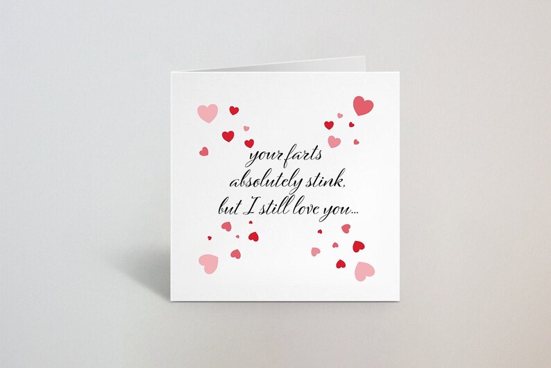 May include: A white greeting card with the text "your farts absolutely stink, but I still love you..." in black script. Red and pink hearts are scattered around the text, creating a romantic and humorous design.