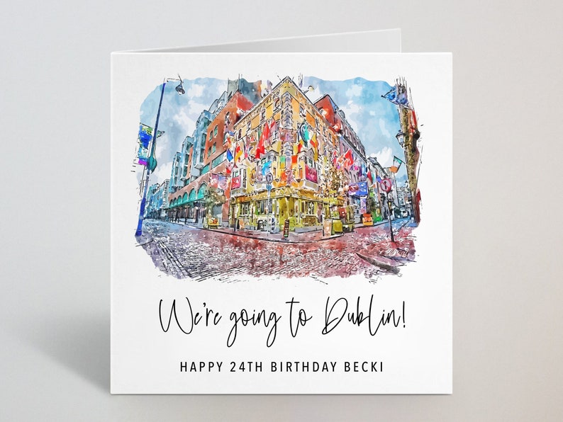 Puede incluir: Tarjeta de felicitaci&oacute;n cuadrada con una pintura de acuarela de una escena callejera en Dubl&iacute;n, Irlanda. La imagen presenta un edificio con banderas y una calle adoquinada. El texto dice "We're going to Dublin!" y "Happy 24th Birthday Becki".