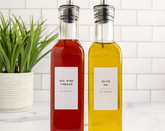 May include: Two glass bottles with pour spouts, one filled with red wine vinegar and the other with olive oil. Both bottles have white labels with black text that reads "Red Wine Vinegar" and "Olive Oil".