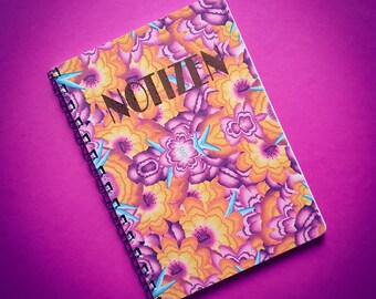 May include: A spiral-bound notebook featuring a colourful floral design in orange, purple, and yellow hues. The word "NOTIZEN" is printed in brown across the cover. The notebook is set against a magenta background.