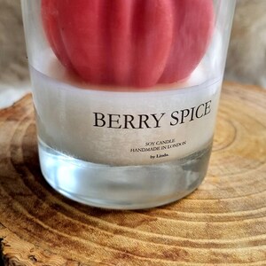 May include: A clear glass candle holder with a red, berry-shaped candle inside. The candle is labeled "Berry Spice" and "Soy Candle Handmade in London by Linda." The candle holder sits on a wooden surface.