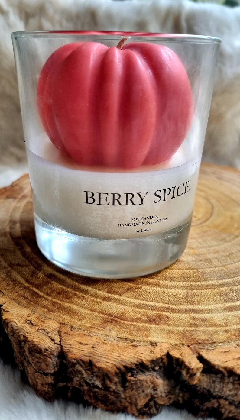 May include: A clear glass candle holder with a red, berry-shaped candle inside. The candle is labeled "Berry Spice" and "Soy Candle Handmade in London by Linda." The candle holder sits on a wooden surface.