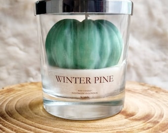 May include: A clear glass candle holder with a silver rim contains a green, pumpkin-shaped candle. The candle is labeled "WINTER PINE" and is handmade in London. The candle sits on a wooden surface.
