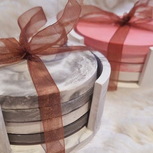 May include: Set of four gray and white marble coasters with a brown ribbon tied around them. The coasters are stacked in a holder.