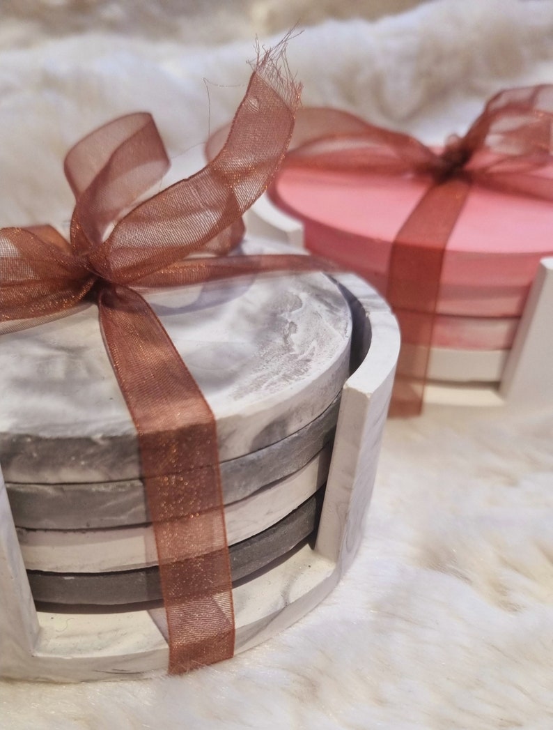 May include: Set of four gray and white marble coasters with a brown ribbon tied around them. The coasters are stacked in a holder.