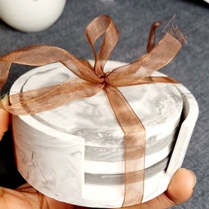 May include: Set of four gray and white marble coasters with a brown ribbon tied around them. The coasters are stacked on top of each other.