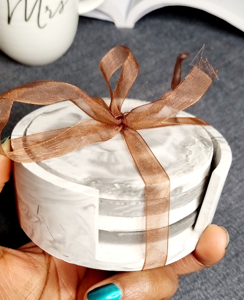 May include: Set of four gray and white marble coasters with a brown ribbon tied around them. The coasters are stacked on top of each other.