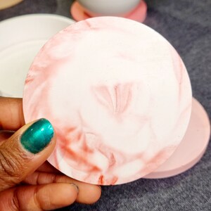 May include: A pink and white marbled round coaster. The coaster is made of resin and has a smooth, glossy finish.