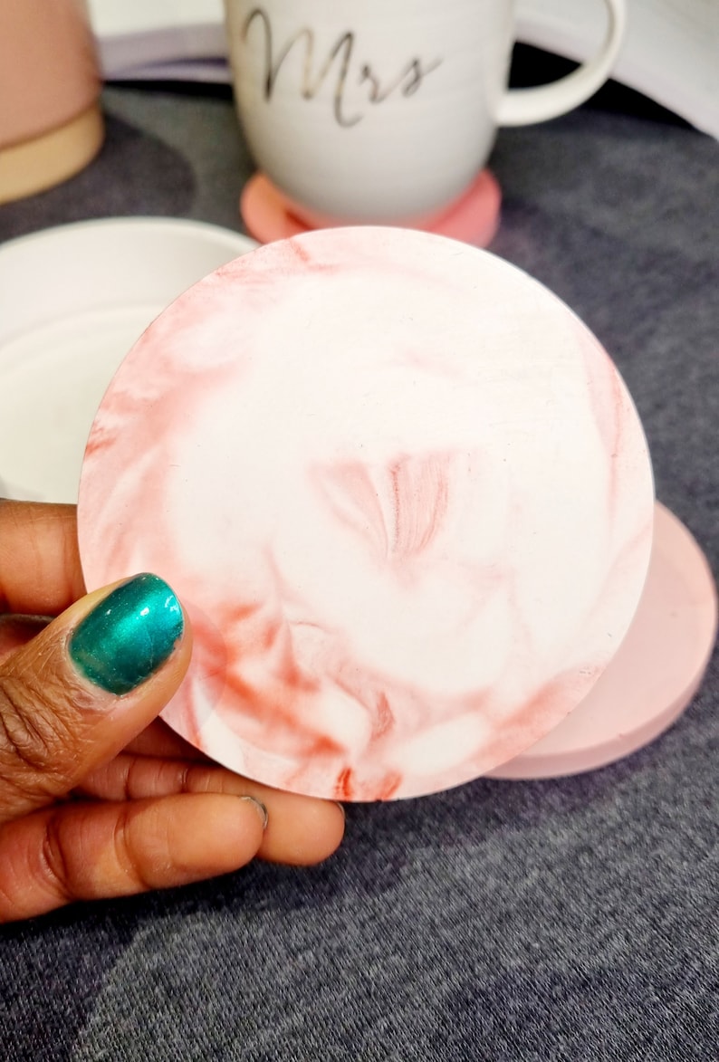 May include: A pink and white marbled round coaster. The coaster is made of resin and has a smooth, glossy finish.