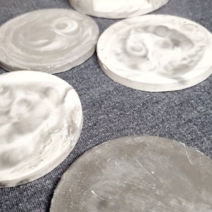 May include: Set of five gray and white concrete coasters. The coasters have a marbled effect and are round. One coaster is taller and has a cutout for a mug.
