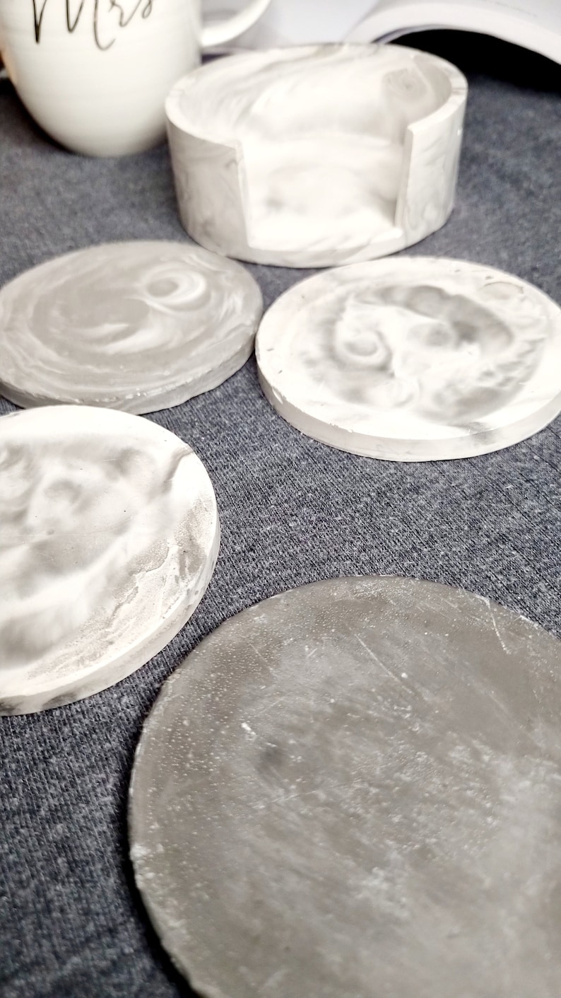 May include: Set of five gray and white concrete coasters. The coasters have a marbled effect and are round. One coaster is taller and has a cutout for a mug.