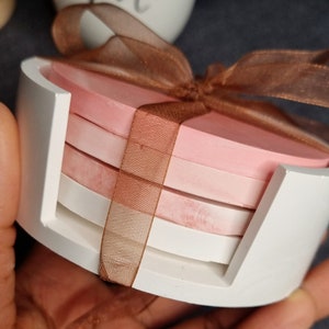 May include: Set of four pink and white coasters with a brown ribbon tied around them. The coasters are in a white holder.