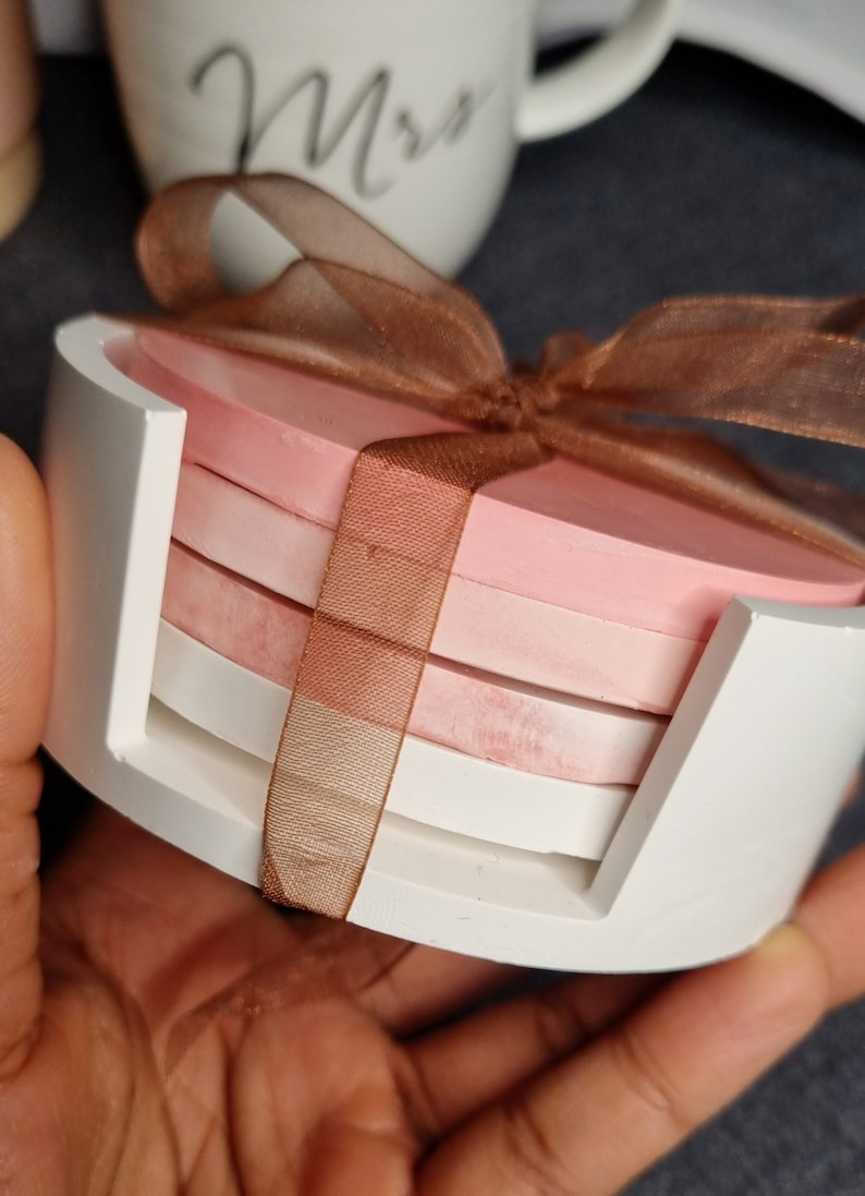 May include: Set of four pink and white coasters with a brown ribbon tied around them. The coasters are in a white holder.