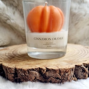 May include: An orange pumpkin-shaped candle in a clear glass. The candle is labeled "Cinnamon Orange" and "Soy Candle Handmade in London". The candle sits on a wooden slice, with a soft, white background.