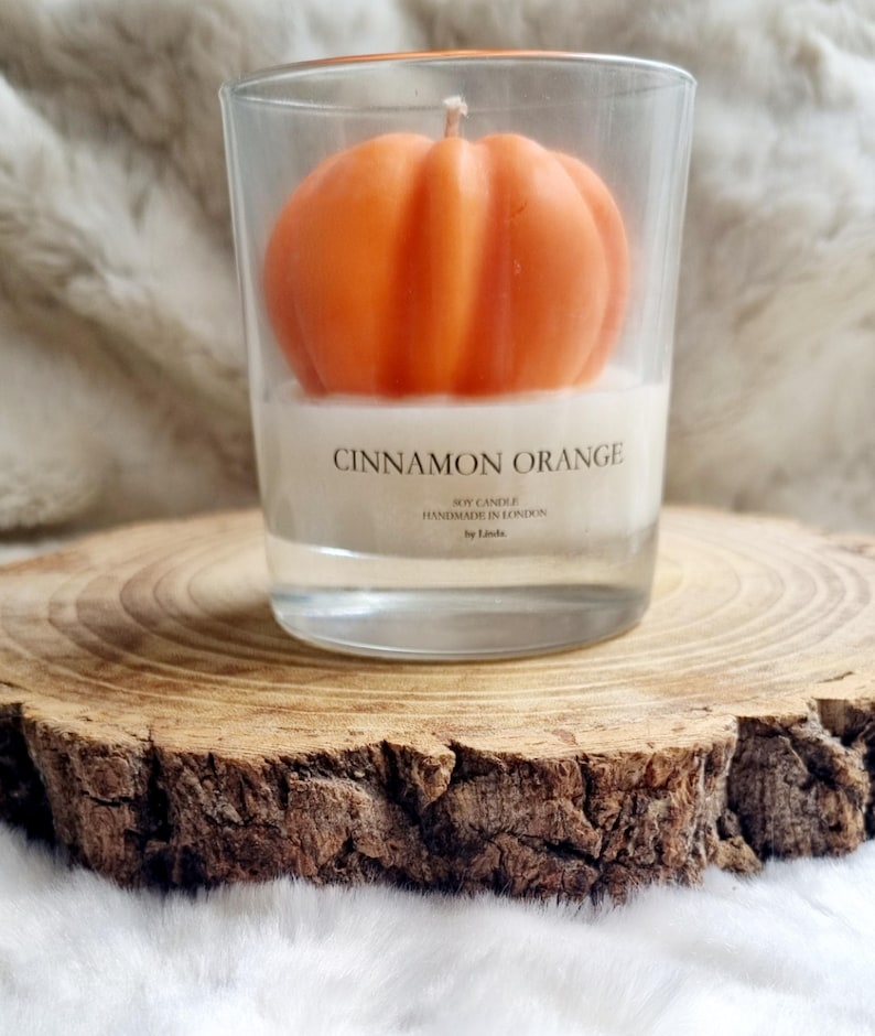 May include: An orange pumpkin-shaped candle in a clear glass. The candle is labeled "Cinnamon Orange" and "Soy Candle Handmade in London". The candle sits on a wooden slice, with a soft, white background.
