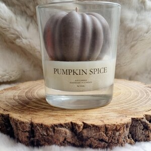 May include: A clear glass candle holder with a pumpkin-shaped candle inside. The candle is a muted brown color and sits atop a white wax base. The label reads "PUMPKIN SPICE". The candle is on a wooden slice.