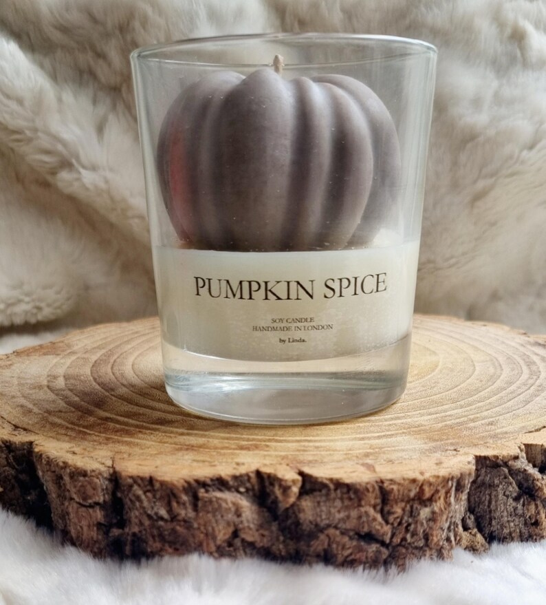 May include: A clear glass candle holder with a pumpkin-shaped candle inside. The candle is a muted brown color and sits atop a white wax base. The label reads "PUMPKIN SPICE". The candle is on a wooden slice.