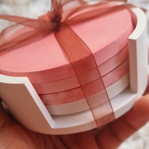 May include: Set of four pink and white round coasters with a brown ribbon tied around them. The coasters are in a white holder.
