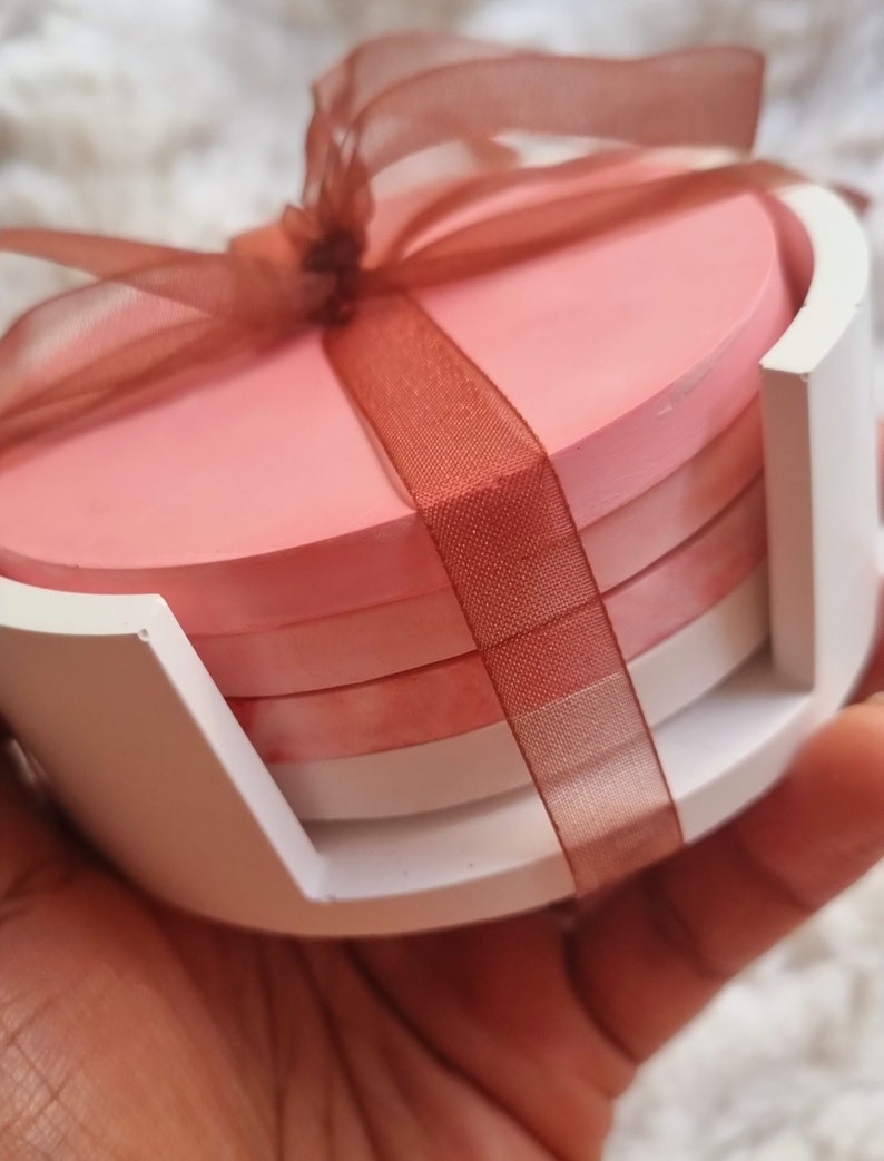 May include: Set of four pink and white round coasters with a brown ribbon tied around them. The coasters are in a white holder.