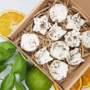 Può includere: Una scatola di carta kraft piena di candele profumate bianche a forma di fiore, cosparse di erbe verdi. La scatola è su una superficie bianca, con fette d'arancia, foglie di basilico verde e un lime. Un prodotto naturale e aromatico.