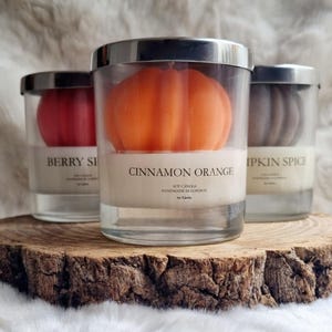 May include: Three soy candles in glass jars with silver lids, each containing a pumpkin-shaped candle. The center candle is orange and labeled "Cinnamon Orange." The other candles are red and brown, labeled "Berry Spice" and "Pumpkin Spice."