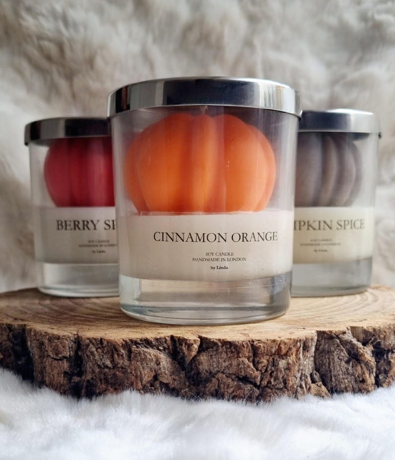 May include: Three soy candles in glass jars with silver lids, each containing a pumpkin-shaped candle. The center candle is orange and labeled "Cinnamon Orange." The other candles are red and brown, labeled "Berry Spice" and "Pumpkin Spice."