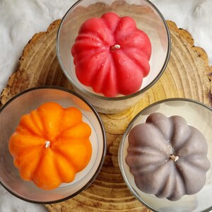May include: Three pumpkin-shaped candles in clear glass holders. The candles are in shades of red, orange, and gray. The candles sit on a wooden slice with a white, fluffy background. Each candle has a white wick.