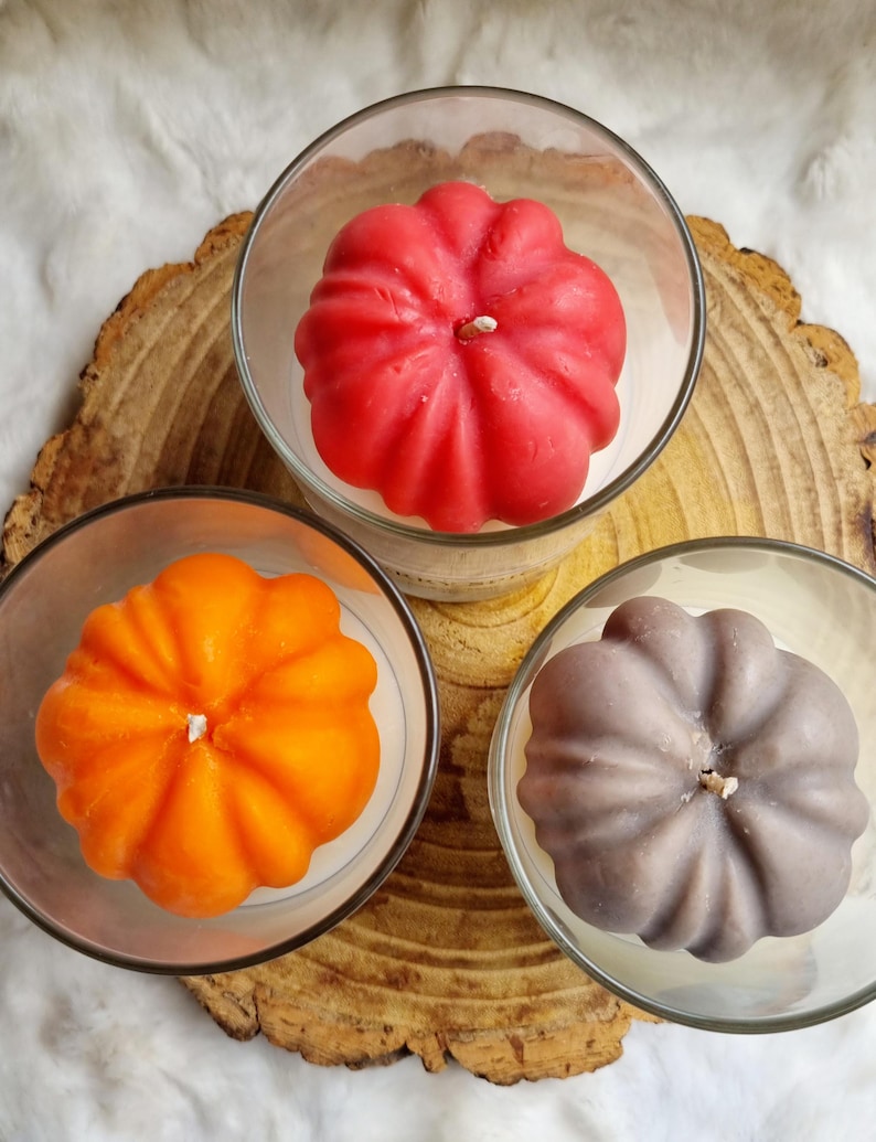 May include: Three pumpkin-shaped candles in clear glass holders. The candles are in shades of red, orange, and gray. The candles sit on a wooden slice with a white, fluffy background. Each candle has a white wick.