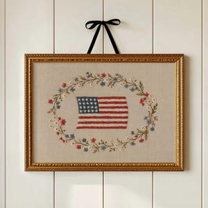 May include: A rectangular framed embroidery piece featuring a central American flag surrounded by an oval wreath of small red, white, and blue flowers. The artwork is encased in a textured gold frame and hangs from a black ribbon bow.