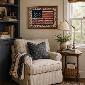 Pode incluir: Uma cena aconchegante de sala de estar com uma poltrona listrada, uma almofada estampada e um cobertor com franjas. Uma obra de arte emoldurada da bandeira americana est&aacute; pendurada acima da cadeira. Uma mesa lateral cont&eacute;m uma l&acirc;mpada, livros e uma pequena cesta.