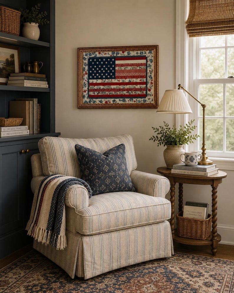 Pode incluir: Uma cena aconchegante de sala de estar com uma poltrona listrada, uma almofada estampada e um cobertor com franjas. Uma obra de arte emoldurada da bandeira americana est&aacute; pendurada acima da cadeira. Uma mesa lateral cont&eacute;m uma l&acirc;mpada, livros e uma pequena cesta.