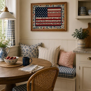 Pode incluir: Um canto de cozinha acolhedor com uma mesa redonda de madeira, uma cadeira de vime e um banco embutido. Na parede, um edredom com a bandeira americana emoldurado. Almofadas e almofadas com padr&otilde;es azuis e brancos.