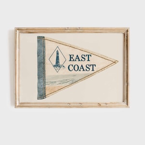 May include: A framed print featuring a triangular pennant flag with a blue denim-textured spine. The off-white pennant displays a blue lighthouse icon inside a diamond shape, the text EAST COAST, and a coastal scene with waves and sand.