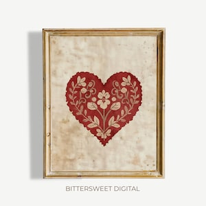 May include: A rectangular wall art piece in a gold frame featuring a deep red heart shape centered on a mottled, aged cream background. The heart contains intricate, light-colored floral and vine patterns. The text BITTERSWEET DIGITAL is visible.