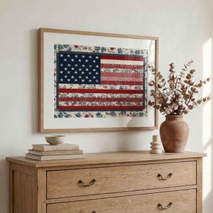 American flag quilt wall art in a light wood frame styled above a wooden dresser with books, vase and neutral farmhouse decor.
